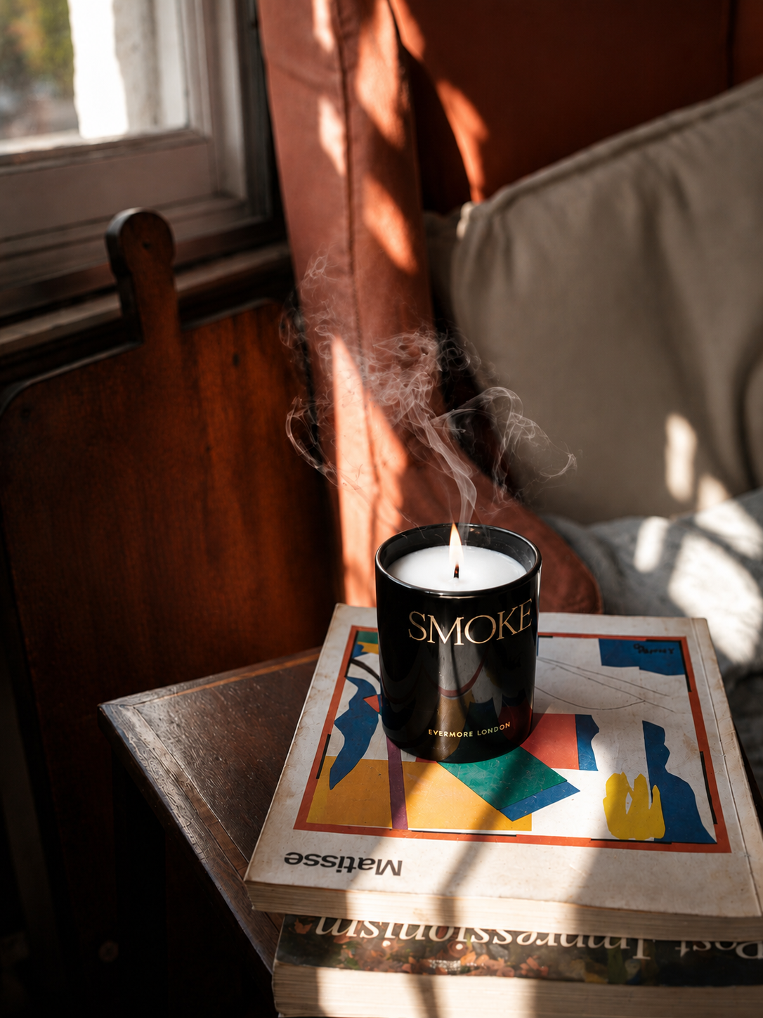 A black scented candle labeled 'SMOKE' sits on top of a stack of books in a sunlit indoor setting, with warm orange-brown curtains, a window, and soft shadows visible around it.